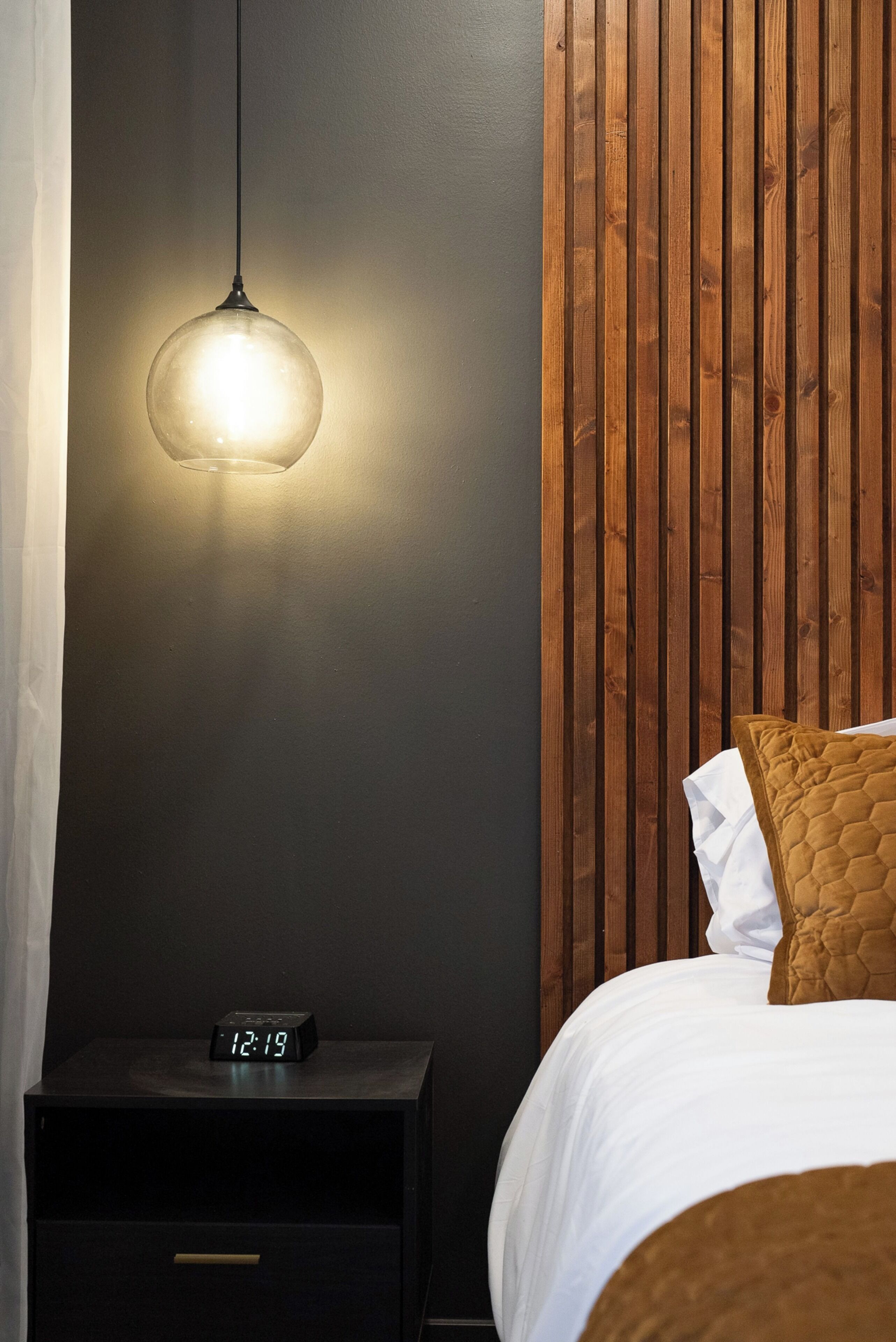 A bedroom with a bed, a nightstand with a digital clock, a hanging light fixture, and a wooden panel wall.