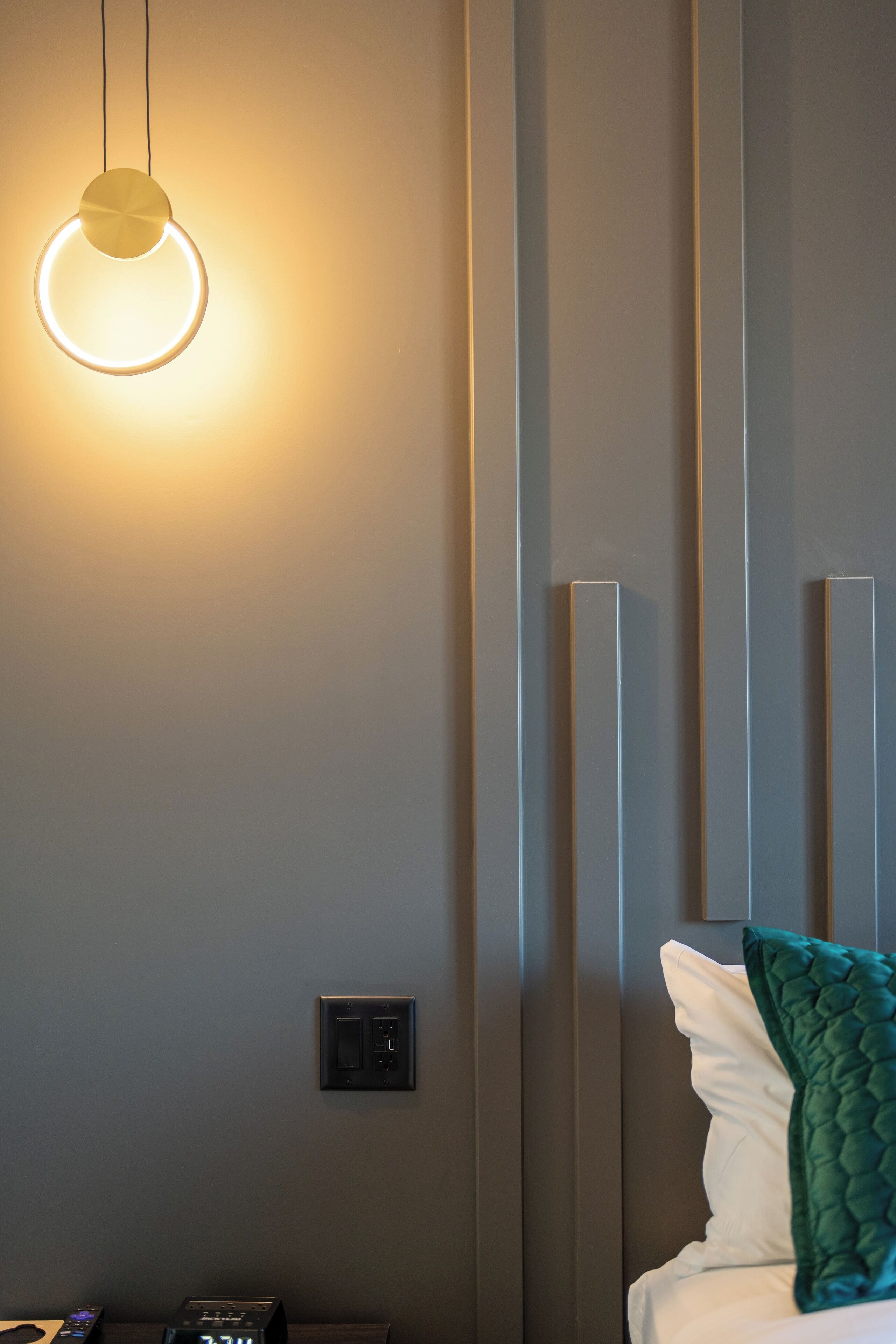 A modern hotel room with a bed, a bedside table with a clock, and a wall-mounted light fixture.