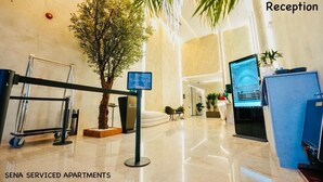 Zona de recepción con mostrador, pantalla digital, un árbol en maceta y un área de descanso.