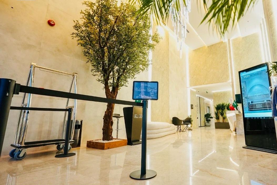 Zona de recepción con mostrador, pantalla digital, un árbol en maceta y un área de descanso.