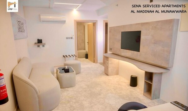 A modern living room with a sofa, ottoman, and a flat-screen TV mounted on the wall. - Sena Serviced Apartments (Madinah)