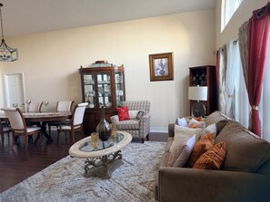 A living room with a dining table, chairs, a glass-top coffee table, a patterned armchair, a sofa, and a cabinet with glass doors. - Charming 2 bedroom house in delightful Naples with pool,WiFi, AC, close to beach (Naples)