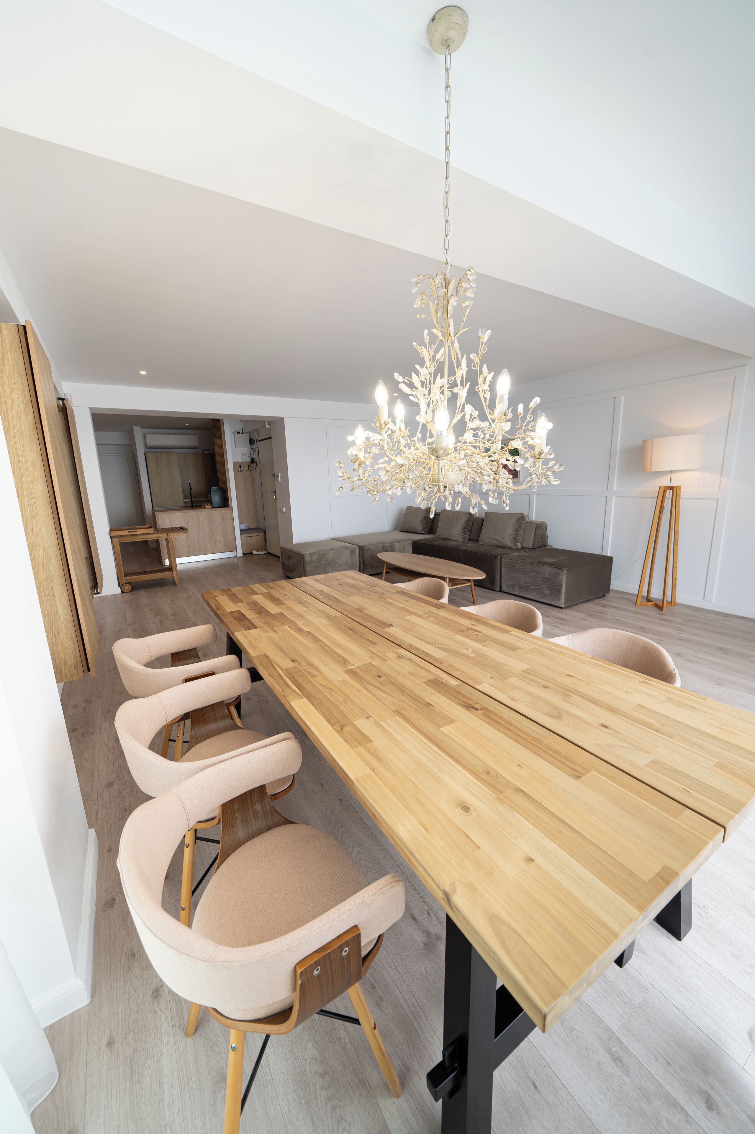Et moderne spiseområde med et langt træbord, polstrede stole, en lysekrone og en grå sofa.