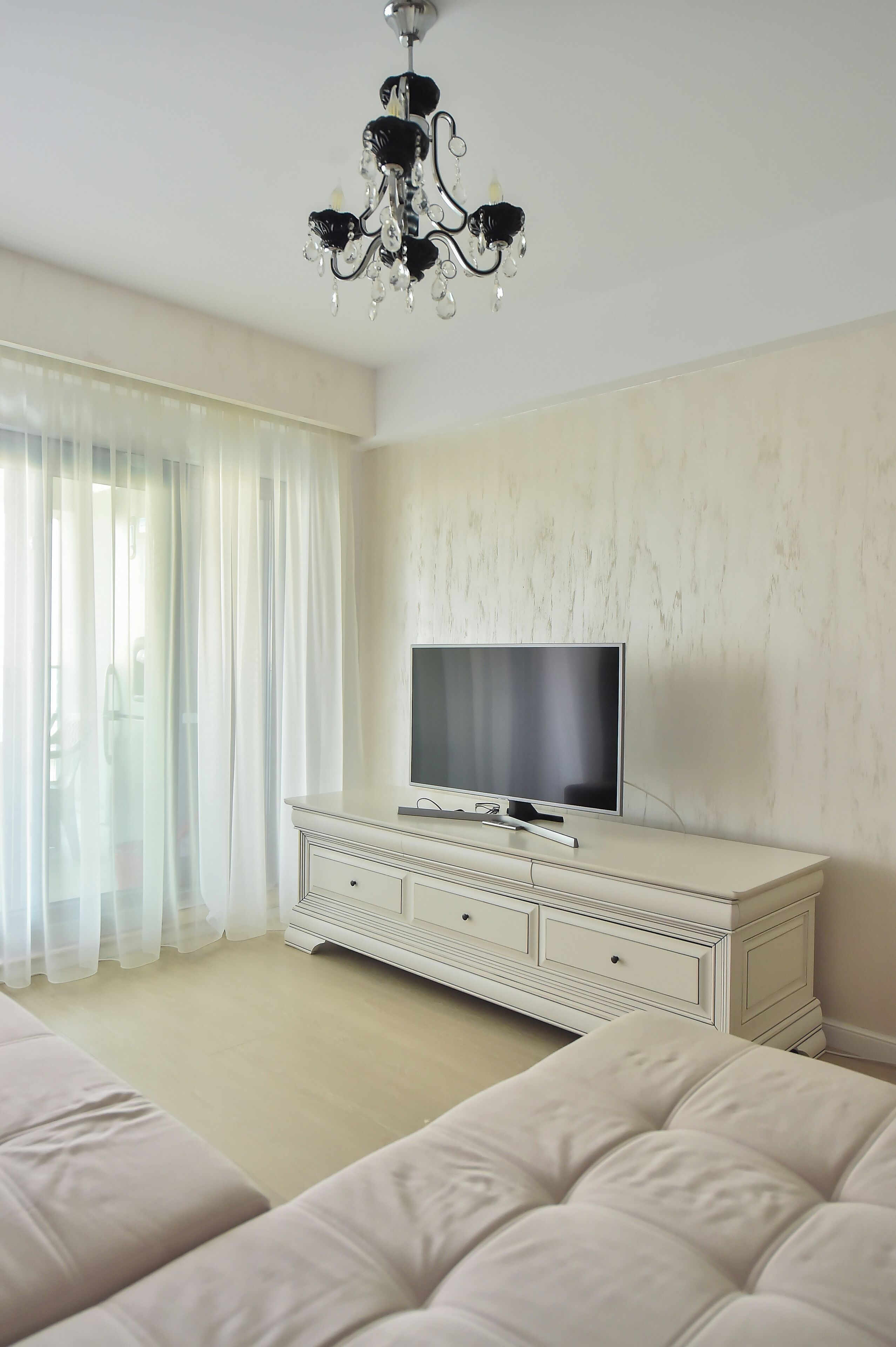 Quarto com televisão sobre uma cômoda branca, um candelabro e uma grande janela com cortinas leves.