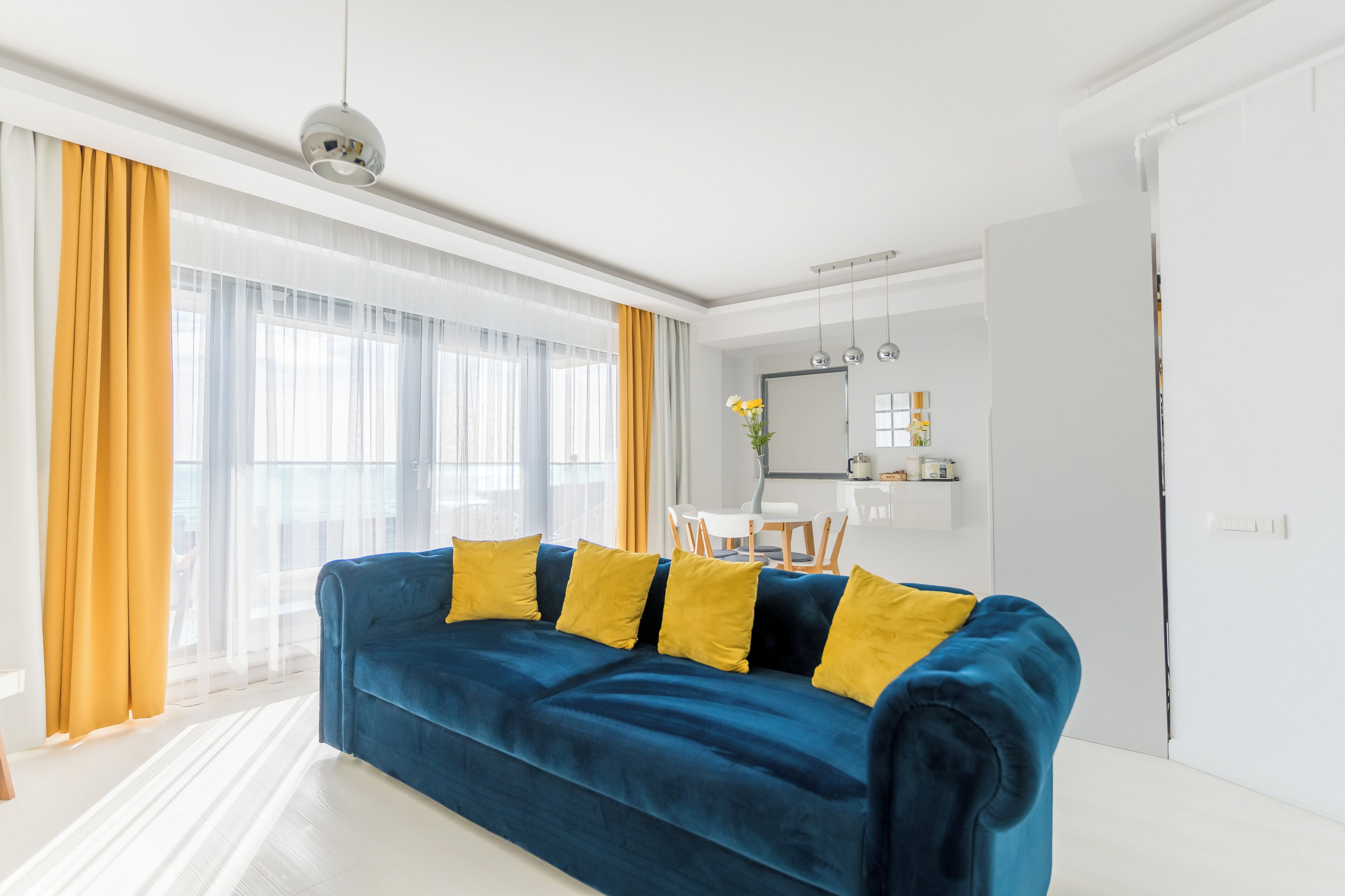 A modern living room with a blue sofa, yellow cushions, and a dining area with a white table and chairs.