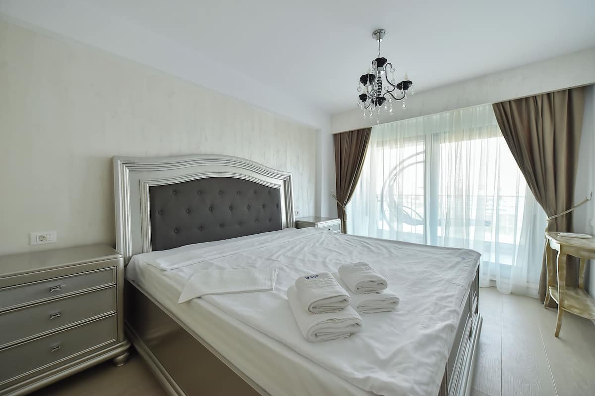A neatly made bed with white linens and towels, a chandelier, a bedside table, and a window with curtains.