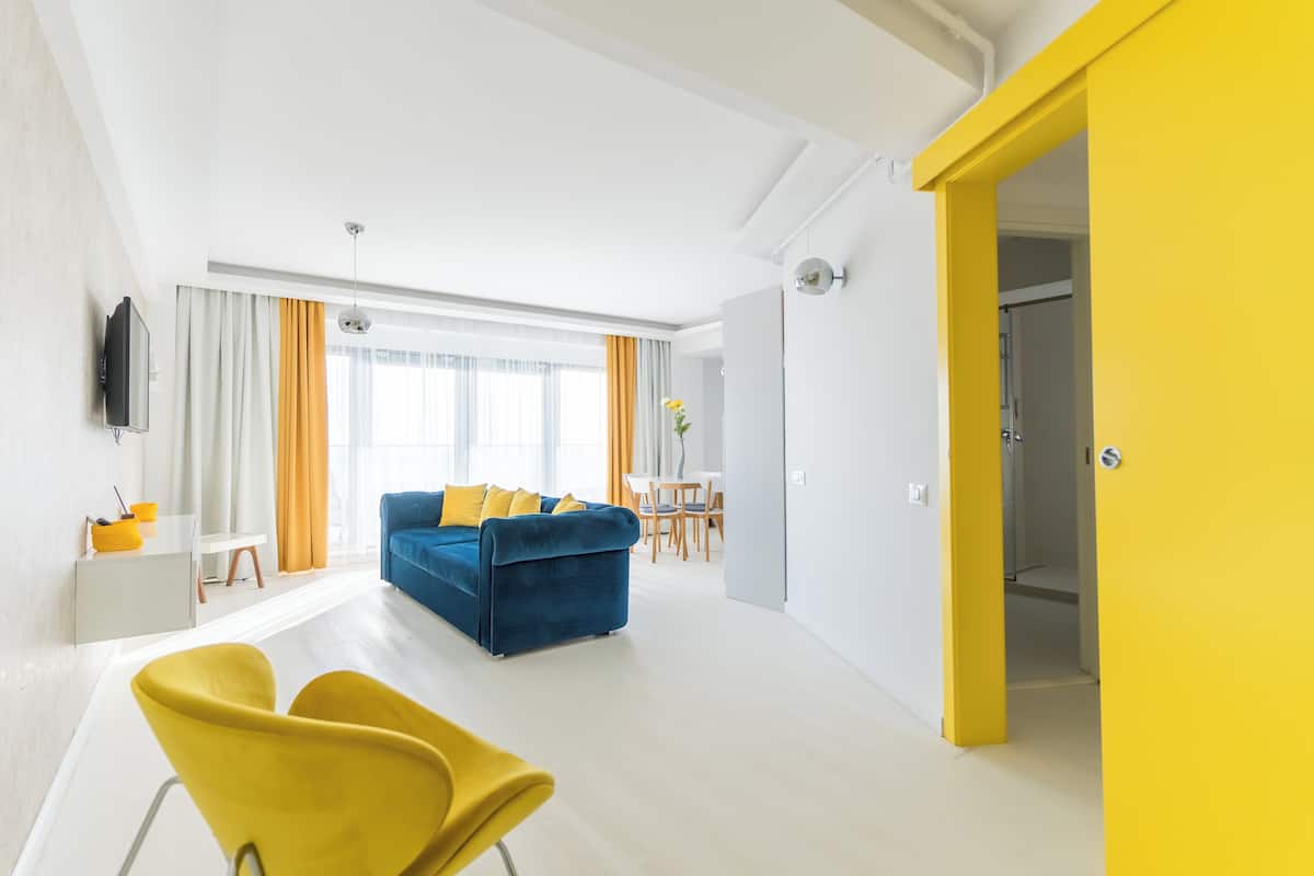 A modern living room with a blue sofa, yellow chairs, a flat-screen TV, and a dining area with a table and chairs.