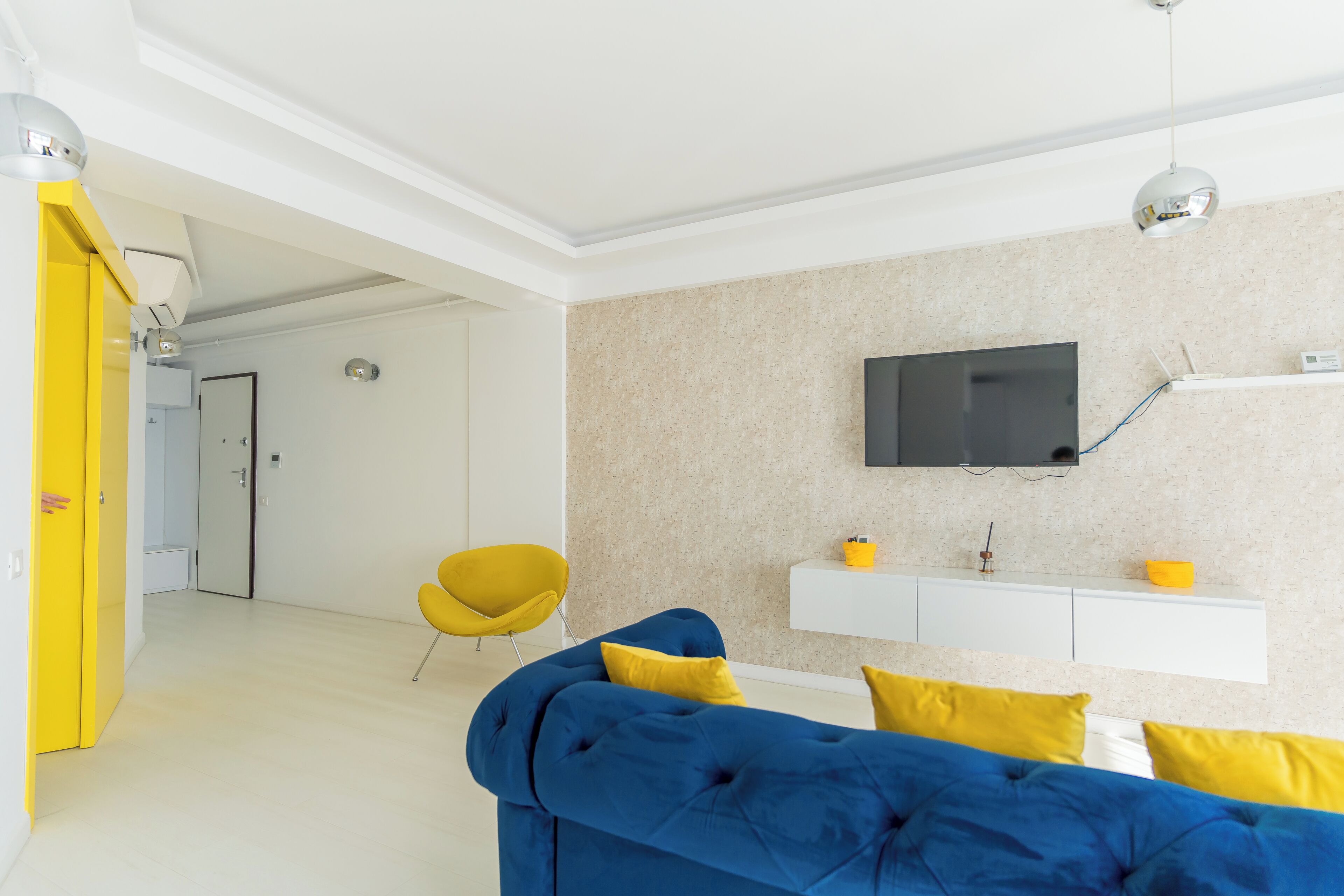 A modern living room with a blue sofa, yellow cushions, a yellow chair, a mounted TV, and a white console table.