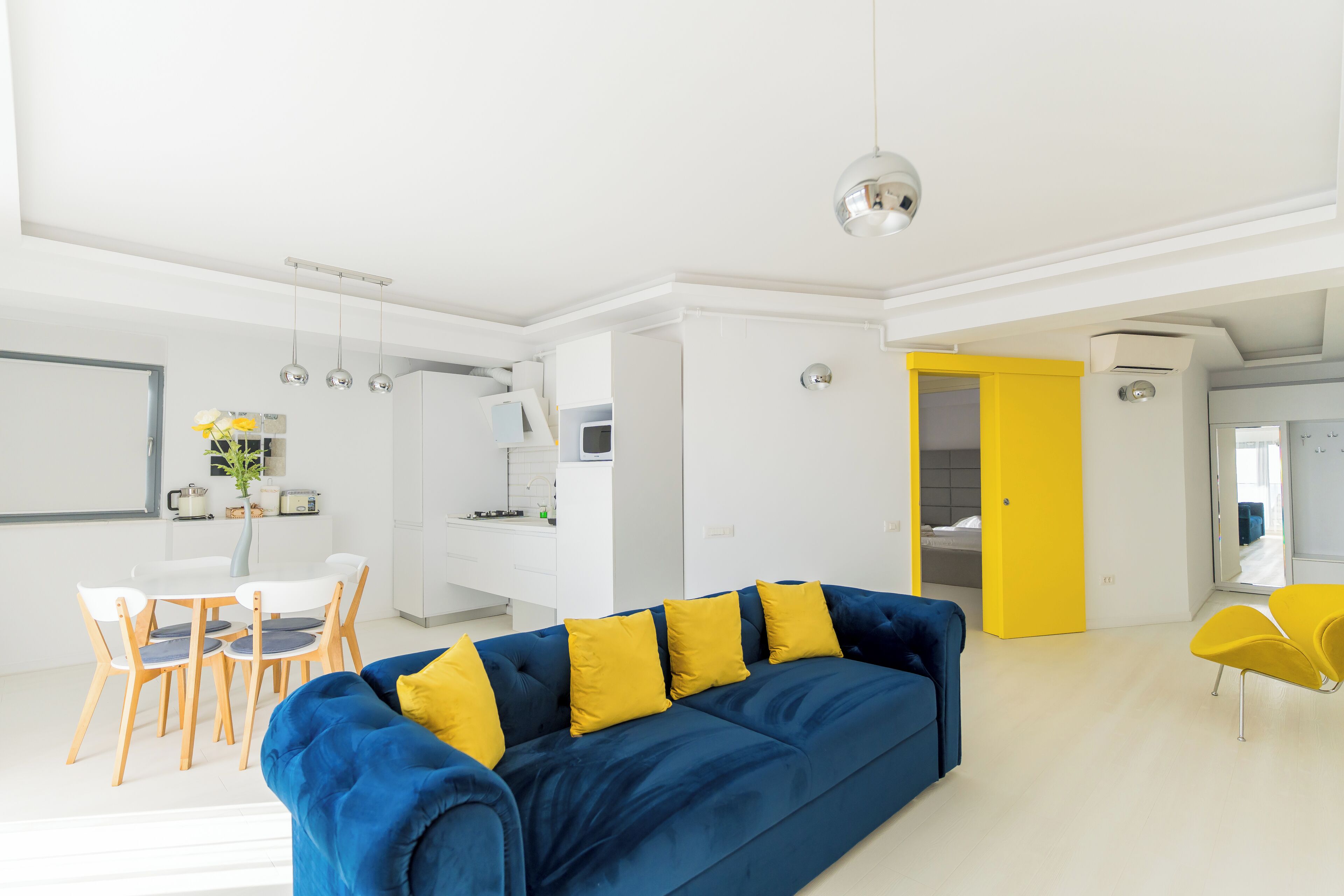 A modern living room with a blue sofa and yellow cushions, a dining area with white chairs, and a bright kitchen in the background.