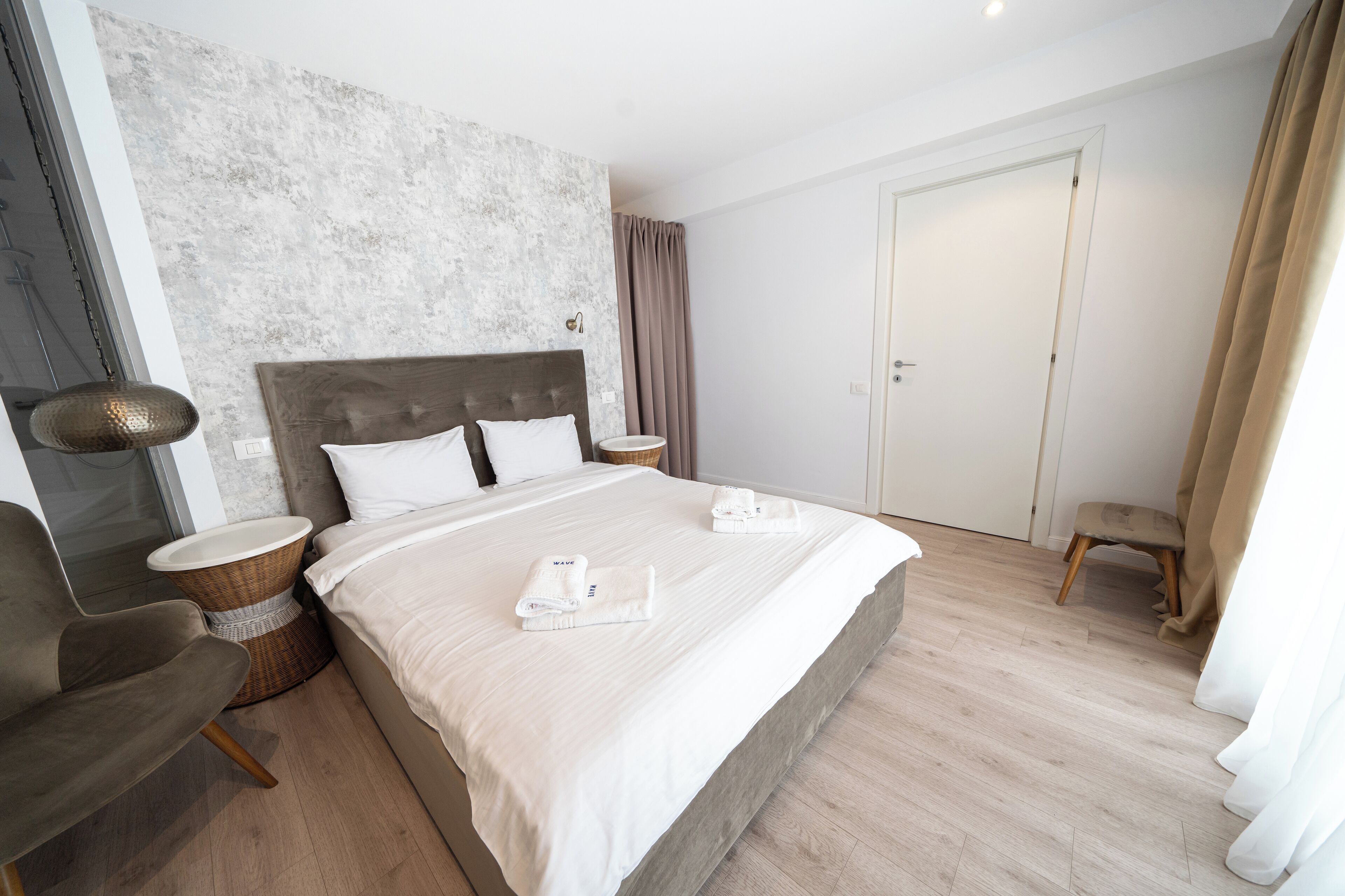Une chambre d’hôtel moderne avec un grand lit, une tête de lit en bois, une table d’appoint en osier, un fauteuil rembourré de couleur grise et un plancher en bois.