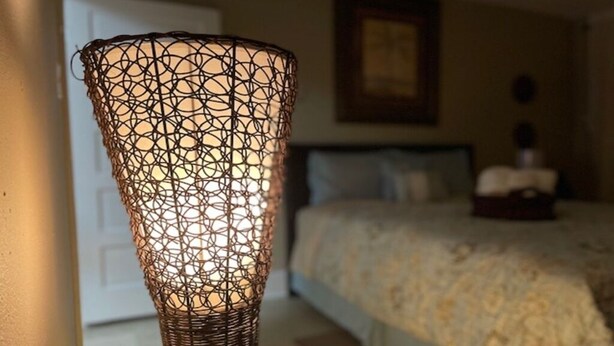 A wicker lamp with a geometric pattern, a bed with a white headboard, and a framed picture on the wall. - Downtown Historic home steps away from Festival International (Lafayette)