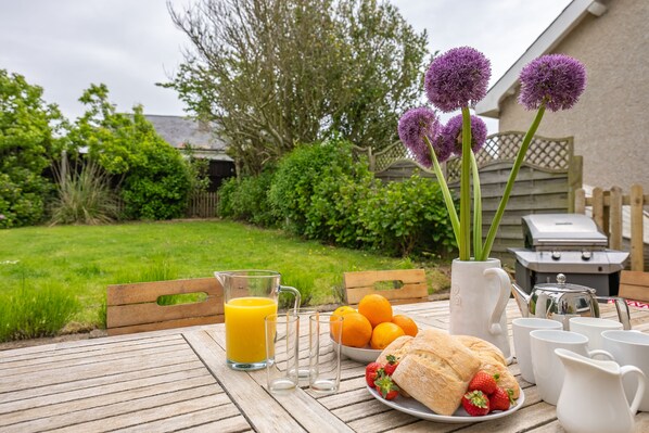 A wooden table with a pitcher of orange juice, a plate of strawberries, a sandwich, a bowl of oranges, and a vase with purple flowers.