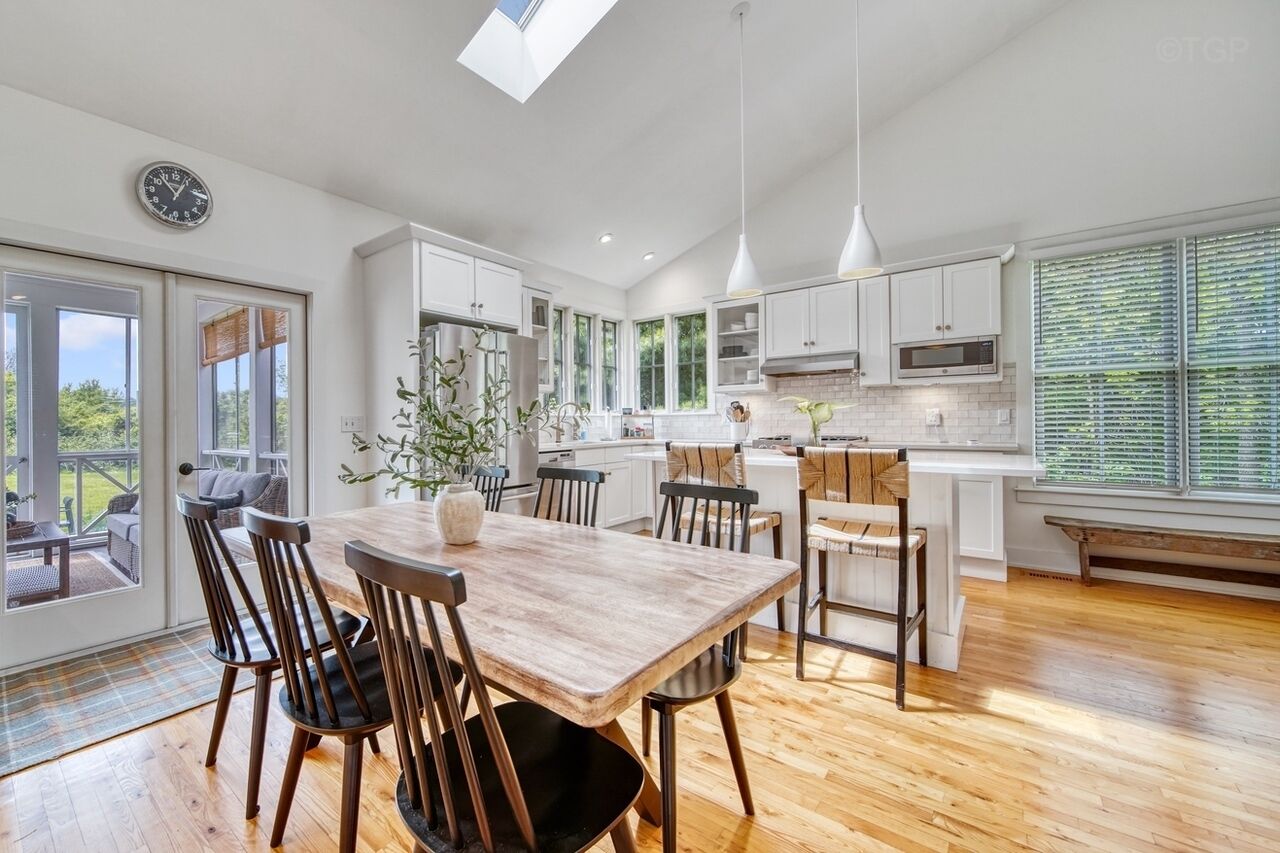 A modern kitchen with white cabinets, a wooden dining table, and a large window with blinds.