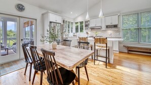 Une cuisine moderne avec des meubles blancs, une table à manger en bois et une grande fenêtre équipée de stores.