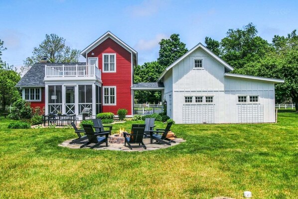 A two-story house with a red and white exterior, a covered porch, and a fire pit in the yard. - Experience tranquility at The Red Farmhouse in secluded Fennville, MI, with 4 bedrooms. (Fennville)
