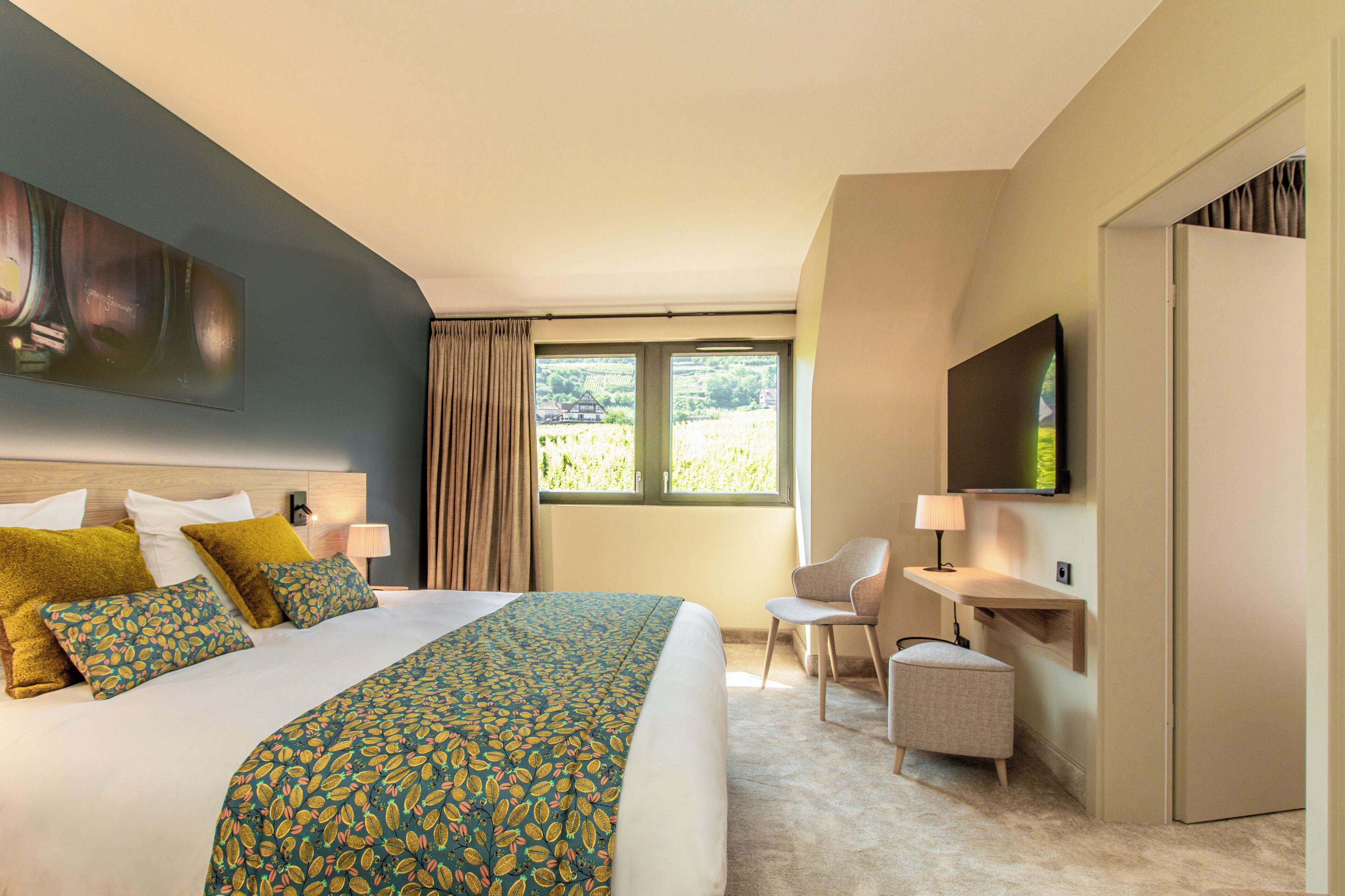 Habitación de hotel con una cama grande, un escritorio, una silla, un televisor y una ventana con vista a la vegetación.