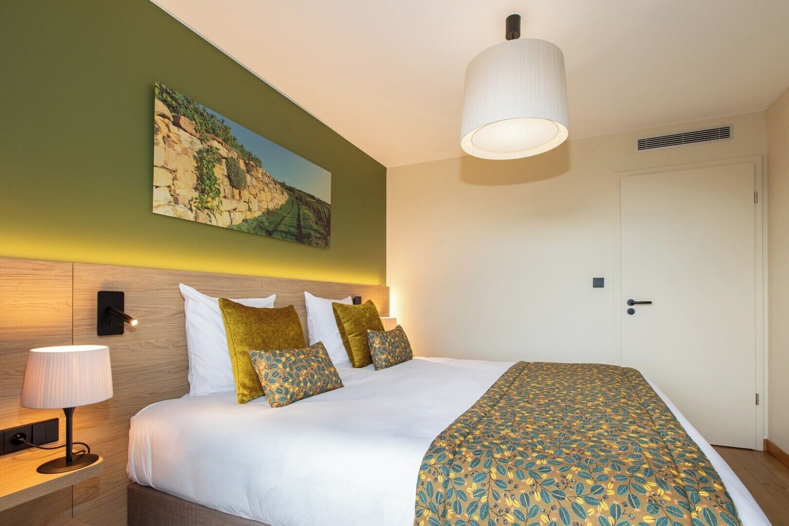 A hotel room with a bed, bedside lamps, a painting on the wall, and a door.