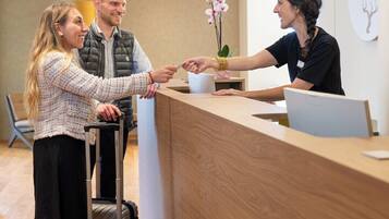 Zona de recepción con un mostrador de madera, una recepcionista y un huésped con una maleta.