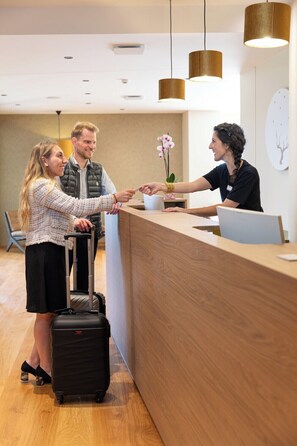 A reception area with a wooden reception desk, a receptionist, and a guest with a suitcase.