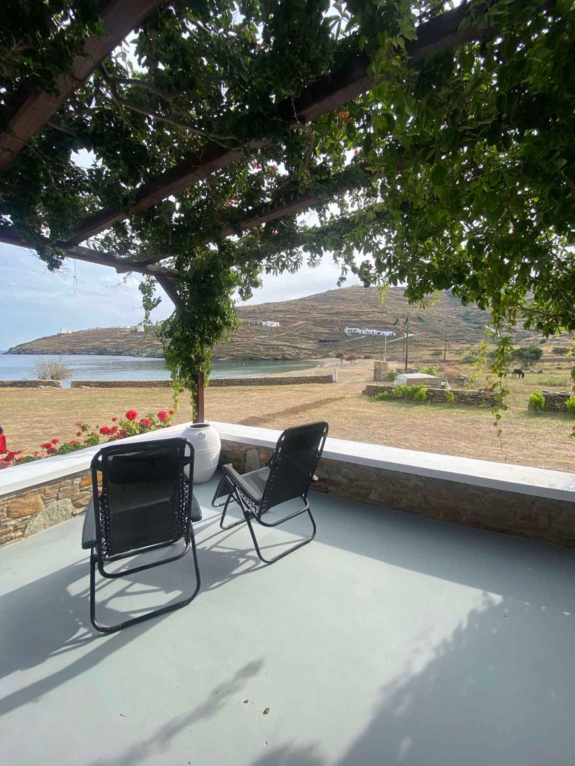 An outdoor seating area with two black metal chairs and a white vase, overlooking a coastal landscape with hills and a body of water.
