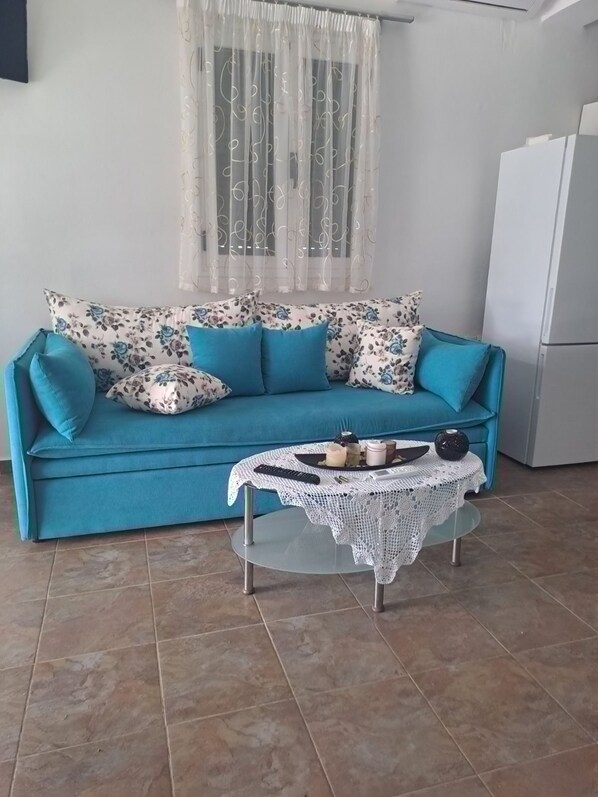 A living room with a teal sofa, floral cushions, a glass coffee table with a white lace table runner, and a white refrigerator in the background.