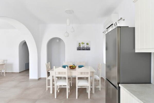 A modern kitchen with white cabinetry, a stainless steel refrigerator, and a dining area with a white table and chairs. - Happy 2-bedroom sea View House Pool Jacuzzi (Paros)