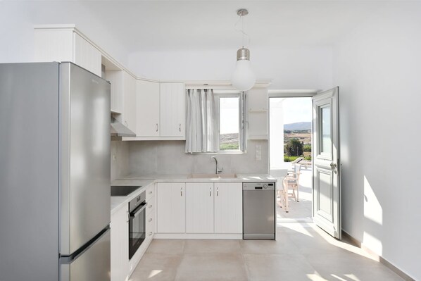 A modern kitchen with white cabinets, a stainless steel refrigerator, a dishwasher, and a window with curtains. - Happy 2-bedroom sea View House Pool Jacuzzi (Paros)