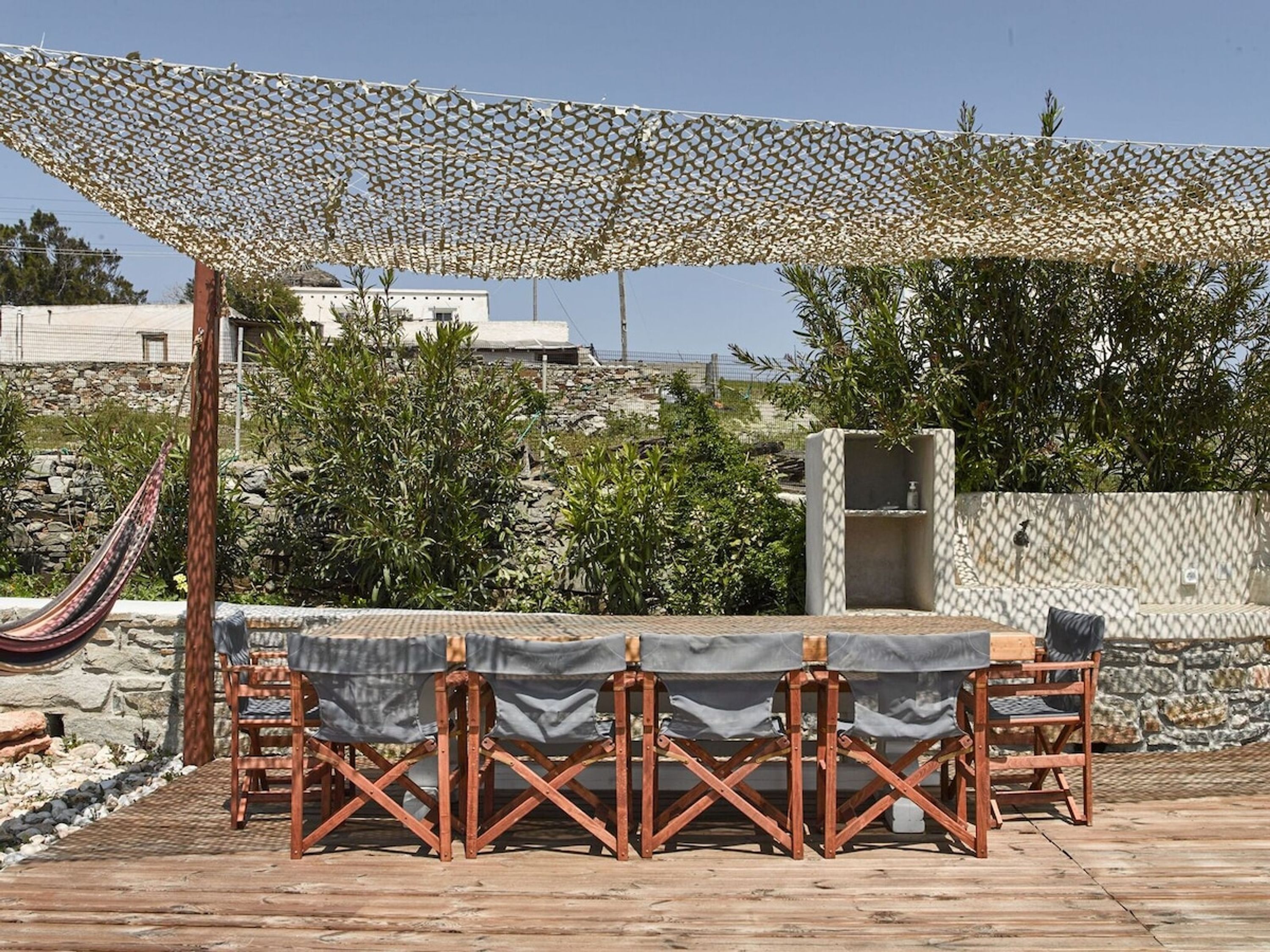 An outdoor patio with a table and chairs under a shaded area, surrounded by plants and a stone wall.