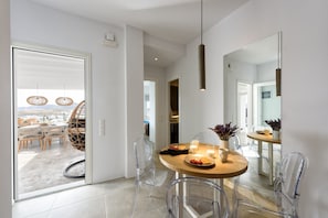 A modern dining area with a round table, clear chairs, and a view through an open door to a balcony with wicker furniture.