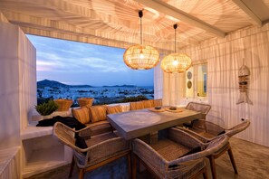 A cozy dining area with wicker furniture, a large window overlooking a scenic landscape, and a ceiling with a unique pattern. - Naousa Story 1 Apartment (Paros)