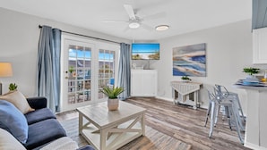 Ein modernes Wohnzimmer mit Sofa, Couchtisch und Fernseher. Eine Glastür öffnet sich zu einem Balkon mit Blick ins Freie.