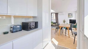 Een moderne keuken met witte kastjes, een broodrooster en een magnetron. Er is een eethoek met een houten tafel en stoelen, en een woonkamer met een televisie en een plant.
