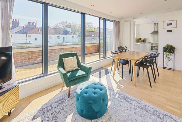 Een moderne woonkamer met een groot raam, een eethoek en een keuken die door het glas zichtbaar is.