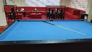 A pool table with a blue felt surface, surrounded by red seating and a large window with a view of a building. - GALAXY-RS-HOTEL (Phnom Penh)