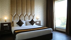 A hotel room with a large bed, two nightstands, a wall-mounted lamp, and a window with curtains. - Hotel Blue Sapphire (Dera Bassi)