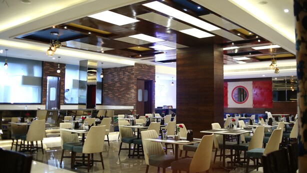 A spacious restaurant with multiple dining tables and chairs, modern ceiling design, and a brick accent wall. - Hotel Blue Sapphire (Dera Bassi)
