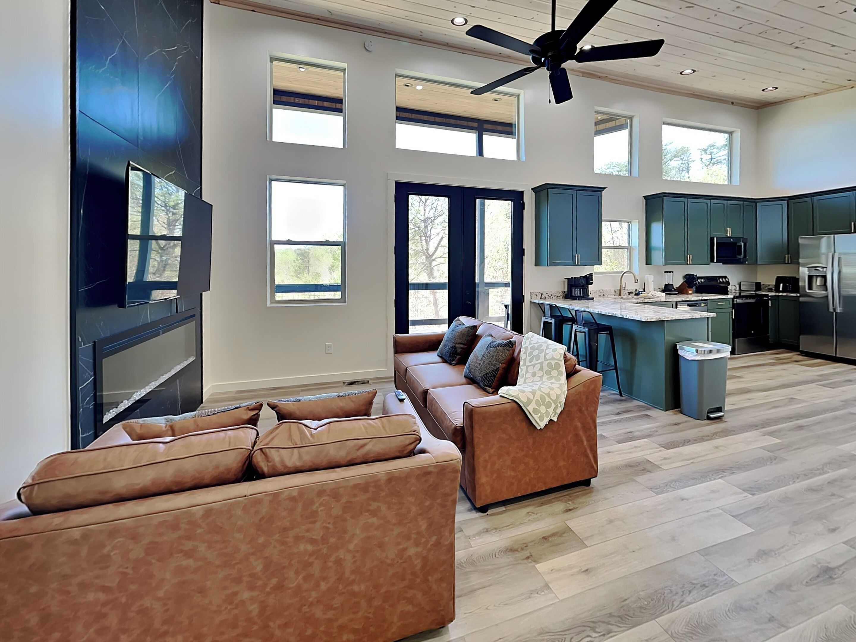 A spacious living room with a brown leather sofa, a ceiling fan, and a kitchen with teal cabinets and modern appliances.