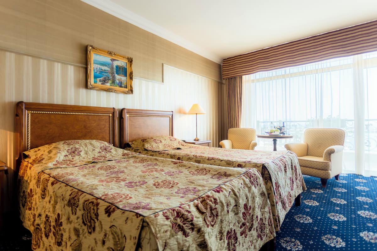 Habitación de hotel con dos camas, un área de descanso con una silla, una mesita y un ventanal con cortinas.