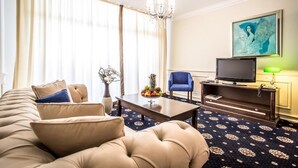 Hotellihuone, jossa on suuri sohva, hedelmillä koristeltu sohvapöytä, televisio ja taulu seinällä.
