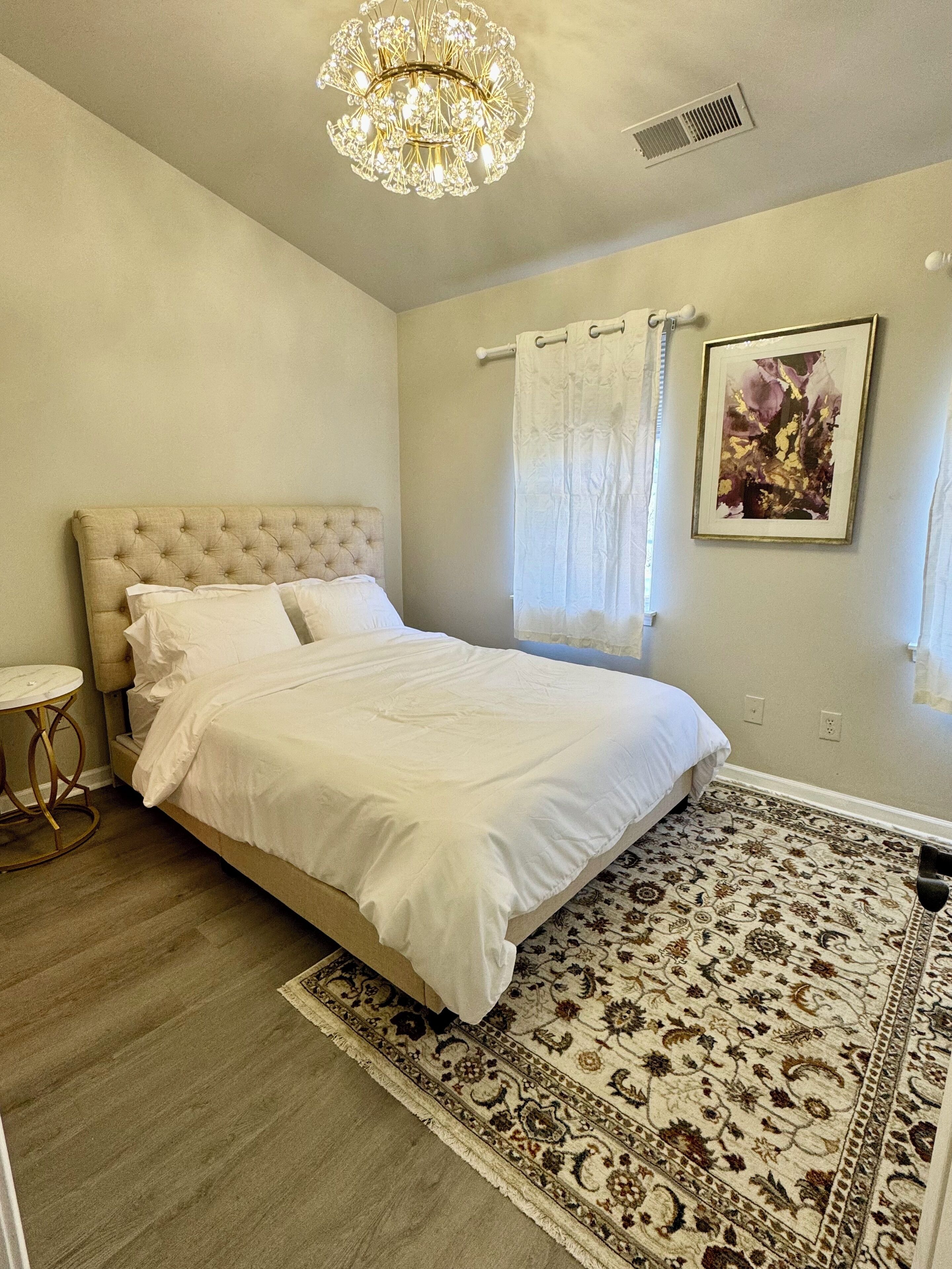 Une chambre à coucher avec un lit doté d’une tête de lit capitonnée, une table de chevet ronde, un tapis à motifs, un lustre et une œuvre d’art encadrée accrochée au mur.