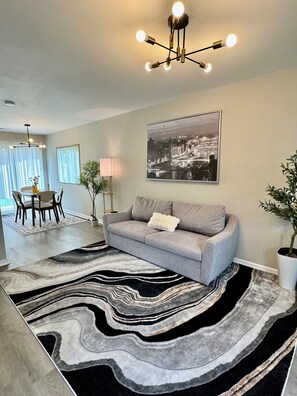Una sala de estar moderna con un sofá gris, un tapete blanco y negro, un cuadro de un paisaje urbano y una zona de comedor al fondo.