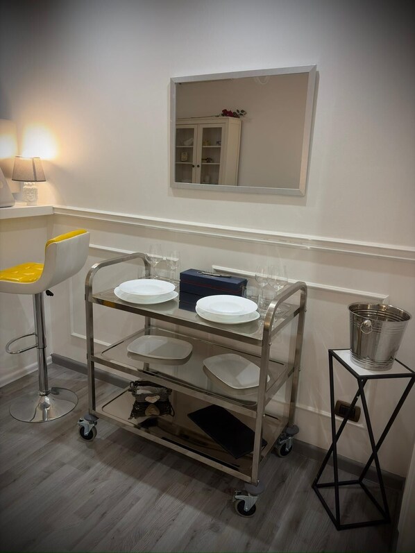 A compact room with a metal cart holding plates and glasses, a small stool with a yellow cushion, a metal bucket on a stand, and a mirror on the wall.