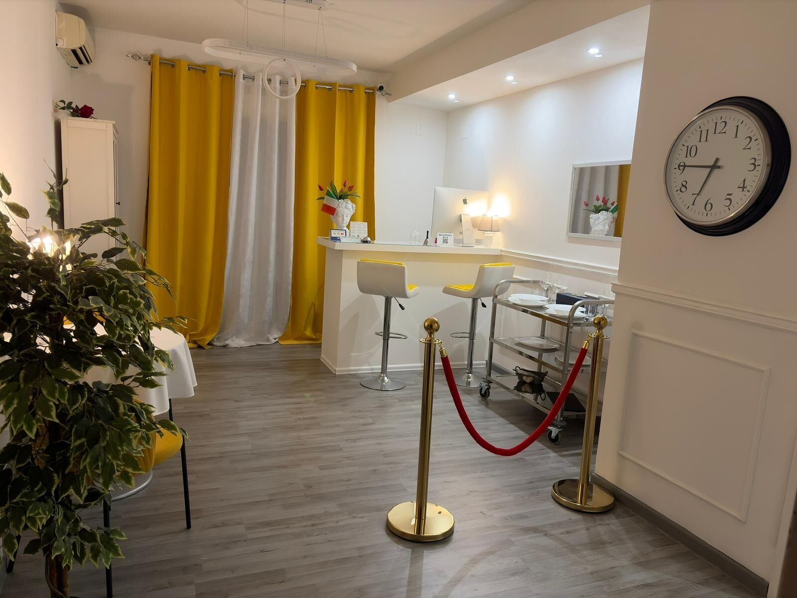 Un espace d’accueil d’hôtel comprenant un comptoir de réception, deux tabourets de bar, une horloge et une plante en pot.