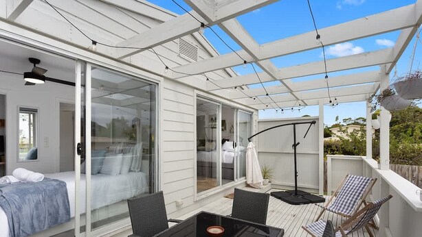 Un patio cubierto con puerta de vidrio que da a un dormitorio, mobiliario exterior y una pérgola con luces colgantes.