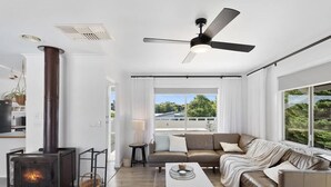 Una sala de estar moderna con chimenea, ventilador de techo y grandes ventanales que ofrecen vistas al verdor exterior.