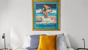 Un cuadro enmarco que muestra a una persona junto al mar, una cama con almohadas y una lámpara sobre una mesita de noche.
