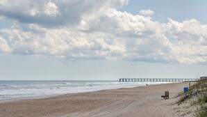 Una playa con un banco de madera, un muelle que se adentra en el océano y un cielo despejado con nubes.