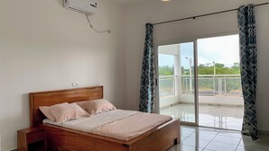 Ein Schlafzimmer mit einem hölzernen Bett, einem Nachttisch, einer Wand-Klimaanlage und einem Balkon mit gemusterten Vorhängen.