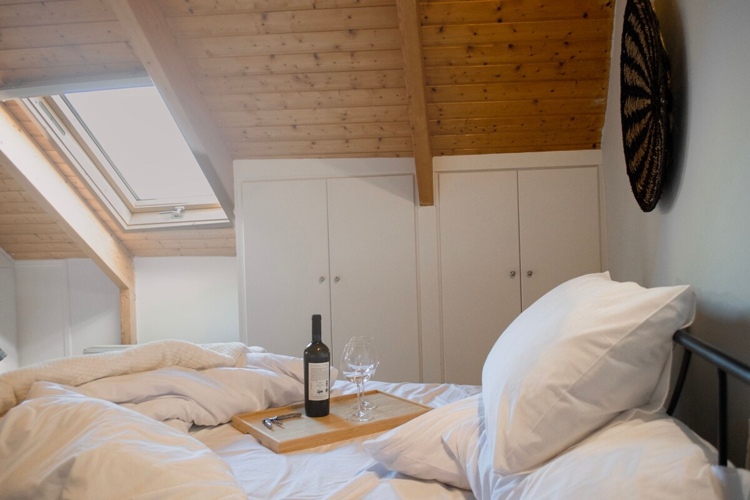 A bedroom with a skylight, wooden ceiling beams, a bed with white bedding, a bedside table with a wine bottle and glass, and a wardrobe.