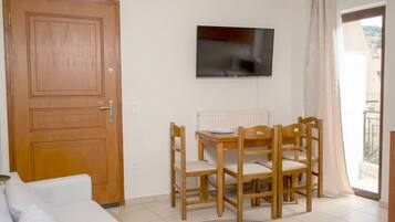 Una habitación con puerta de madera, un sofá blanco, una mesa de madera con sillas, un televisor montado en la pared y vistas a través de una ventana con cortina.